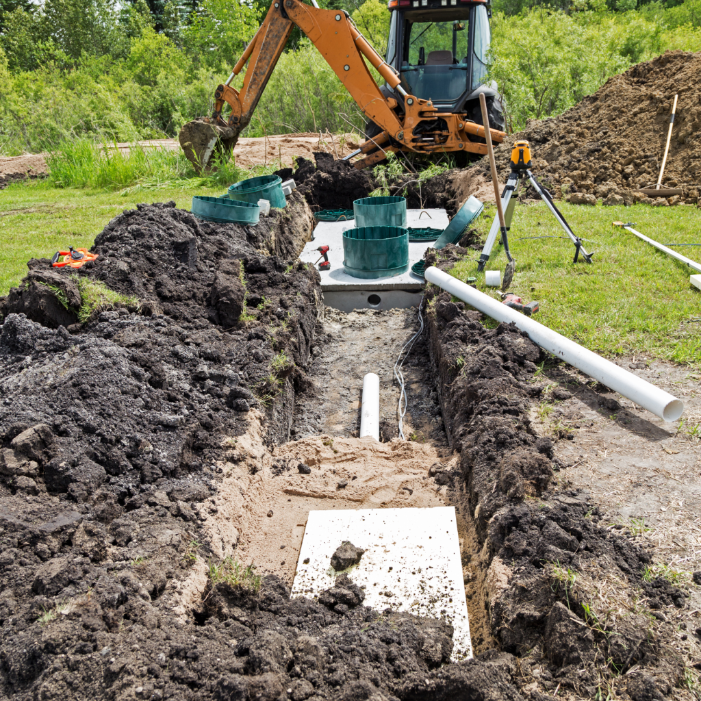 Septic tank installation in progress — concrete tank set in trench with green risers and PVC inlet pipe, excavator and surveying equipment on site